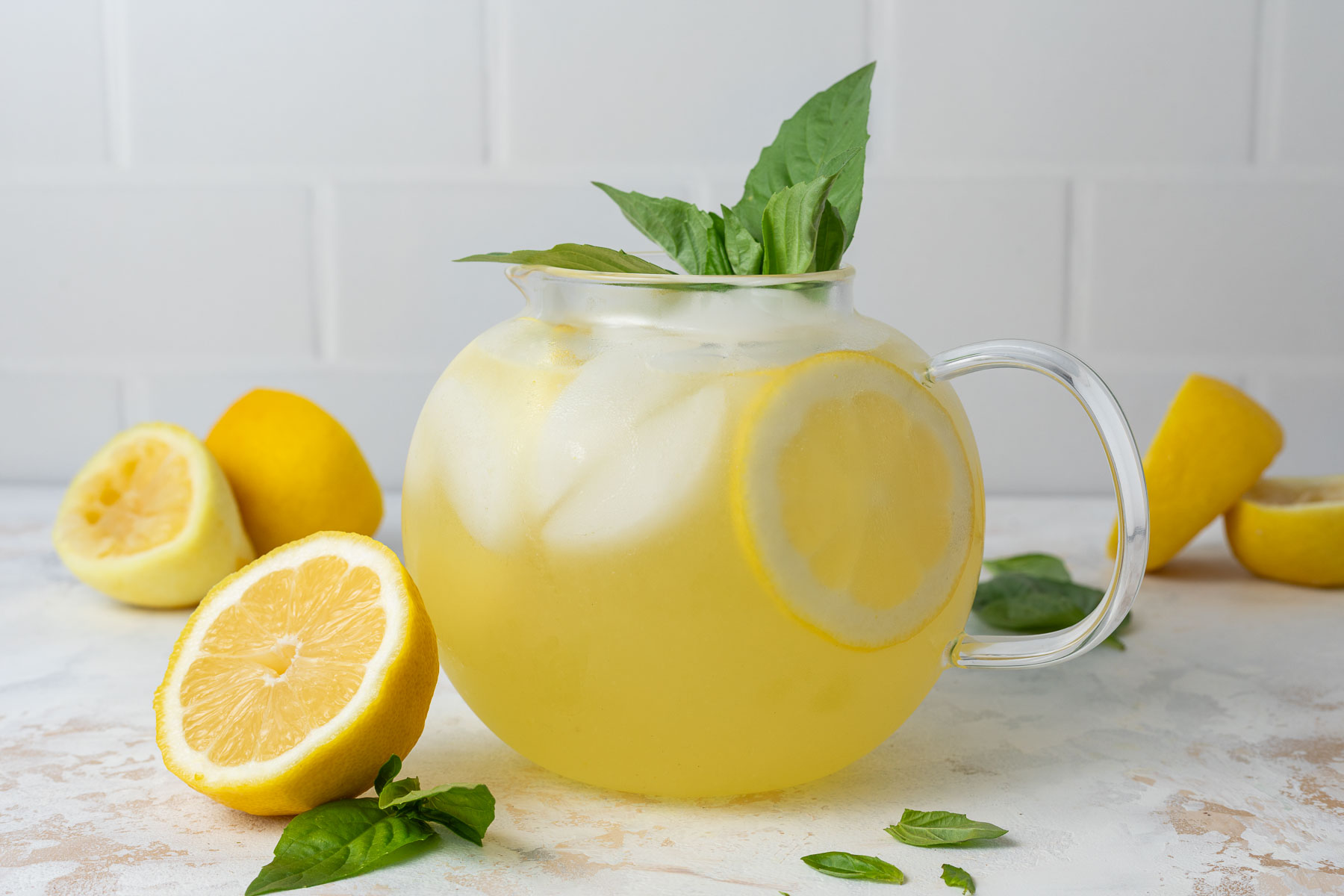 A pitcher of basil lemonade garnished with fresh lemon slices and fresh basil, surrounded by lemon halves and more fresh basil.