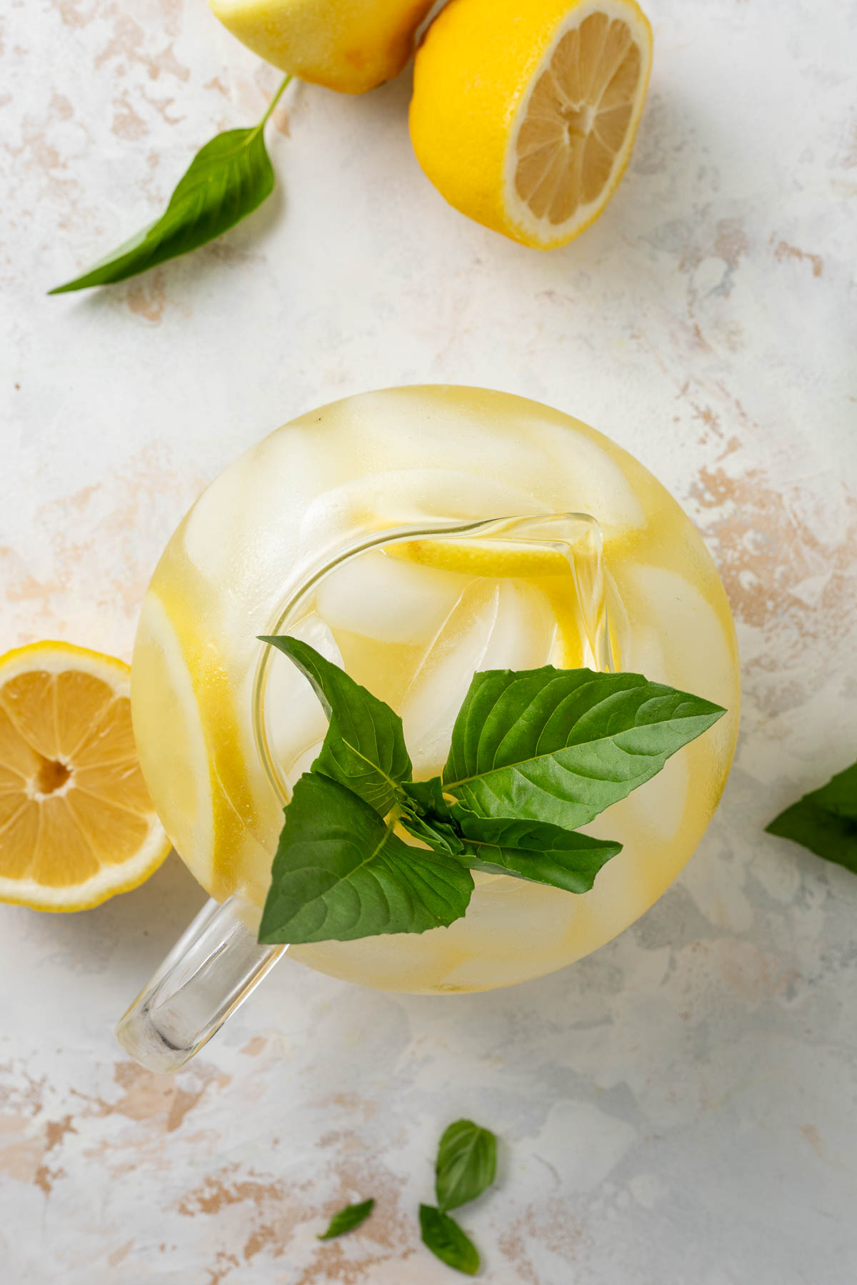 A pitcher of basil lemonade garnished with fresh lemon slices and fresh basil, surrounded by lemon halves and more fresh basil.