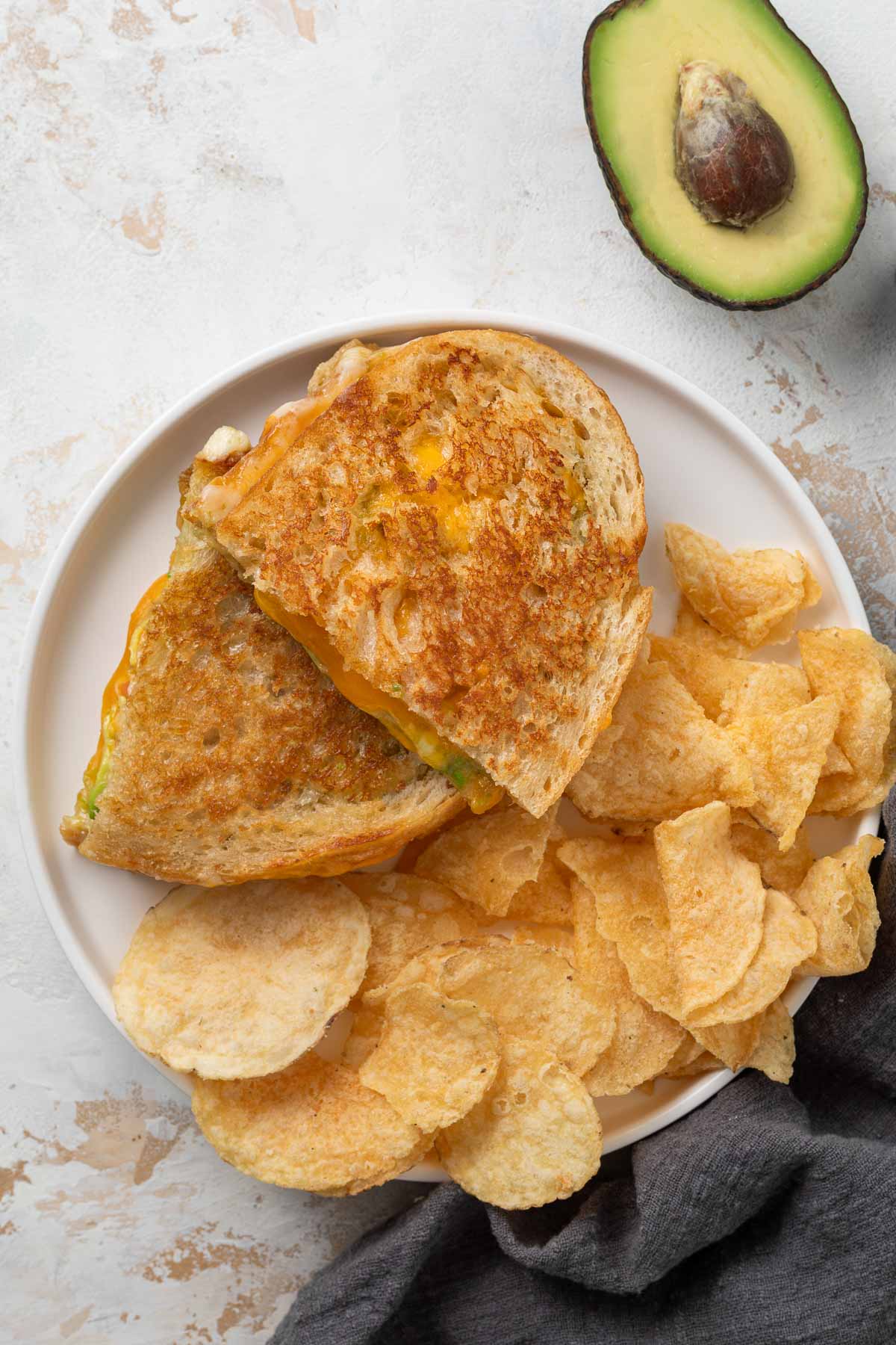 An avocado grilled cheese cut half on a plate with a pile of chips.