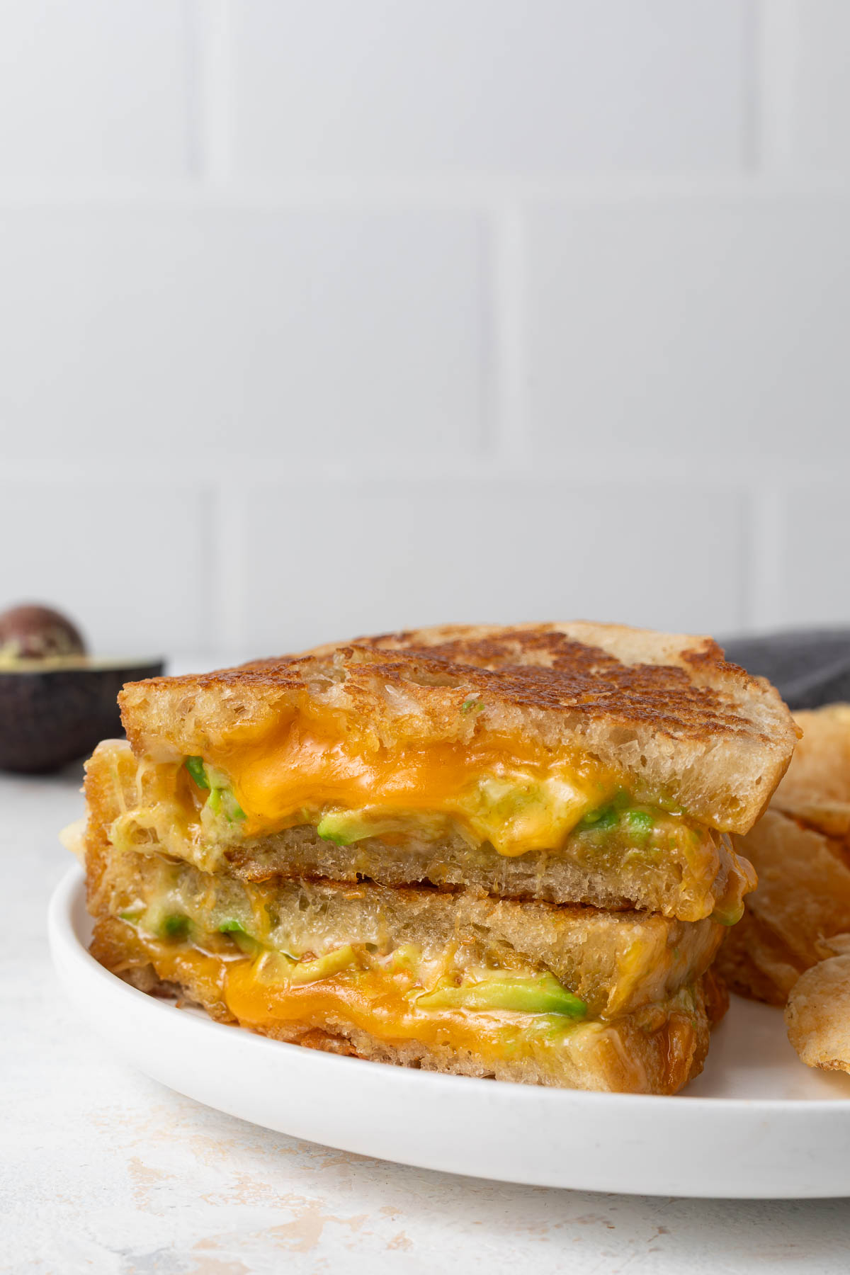 An avocado grilled cheese cut in half revealing a gooey inside. It's a on a plate with a pile of chips.