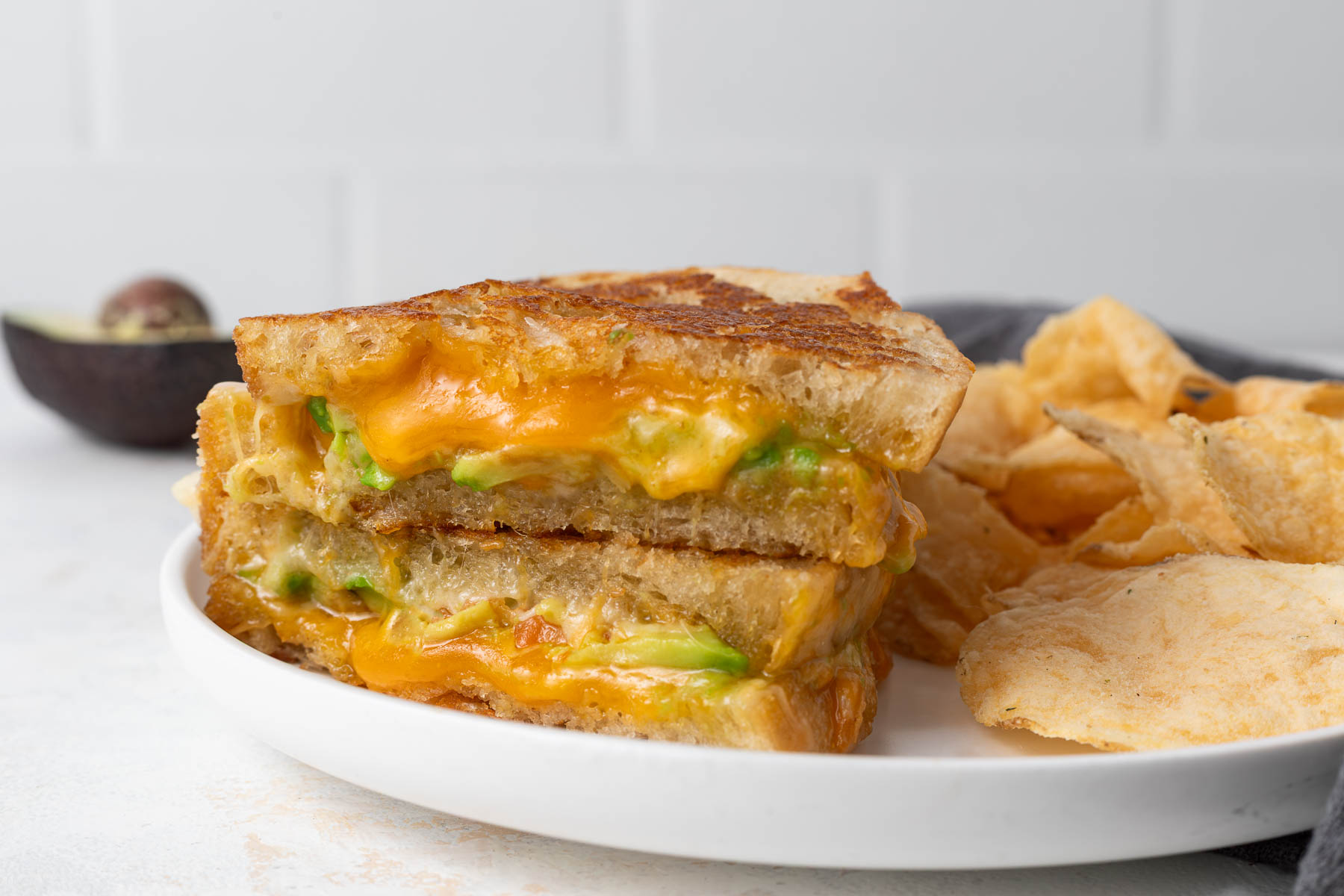 An avocado grilled cheese cut in half revealing a gooey inside. It's a on a plate with a pile of chips.