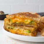 An avocado grilled cheese cut in half revealing a gooey inside. It's a on a plate with a pile of chips.