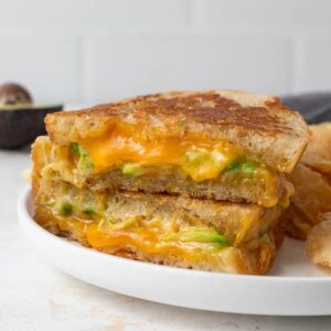 An avocado grilled cheese cut in half revealing a gooey inside. It's a on a plate with a pile of chips.