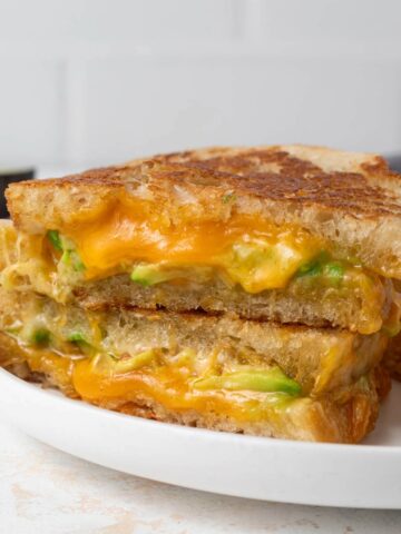 An avocado grilled cheese cut in half revealing a gooey inside. It's a on a plate with a pile of chips.